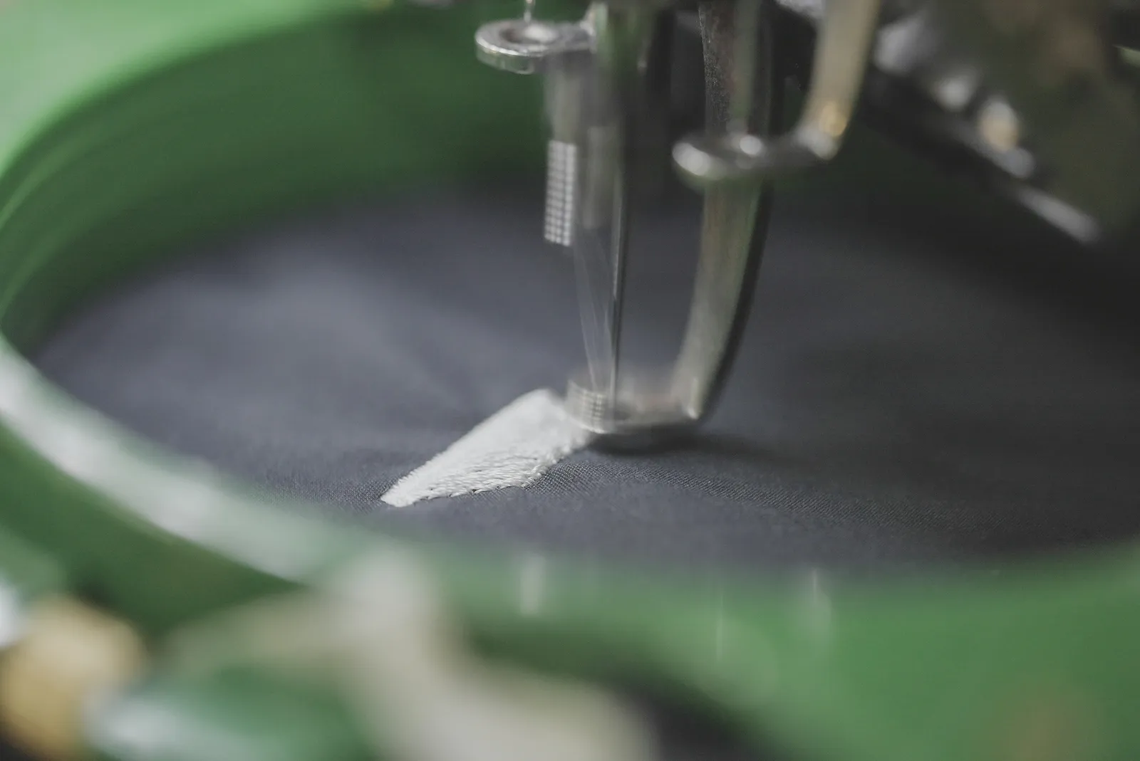 Nahaufnahme: Die Nadel einer Stickmaschine stickt weißes Garn auf dunklen Stoff im Stickrahmen.